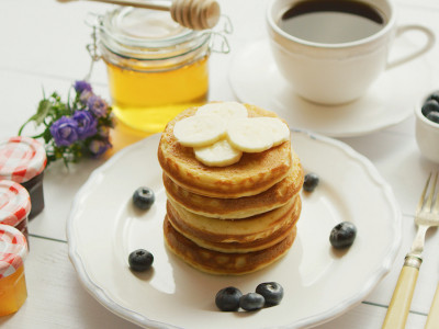 From above view of pancakes served in white plate decorated with slices of banana and blueberries with cup of coffee and honey placed near on white background.